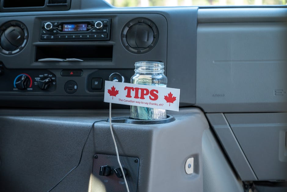 A close-up view inside a vehicle showing a dashboard with various control knobs and a built-in radio. In the foreground, a transparent glass jar filled with coins is placed in one of the cup holders. 